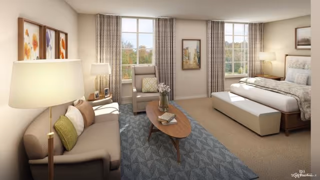 A bright and cozy bedroom with a large bed, a cushioned bench at the foot of the bed, a sofa with pillows, a wooden coffee table with books and a vase of flowers, an armchair, two large windows with curtains letting in natural light, framed artwork on the walls, and two lamps providing warm lighting.