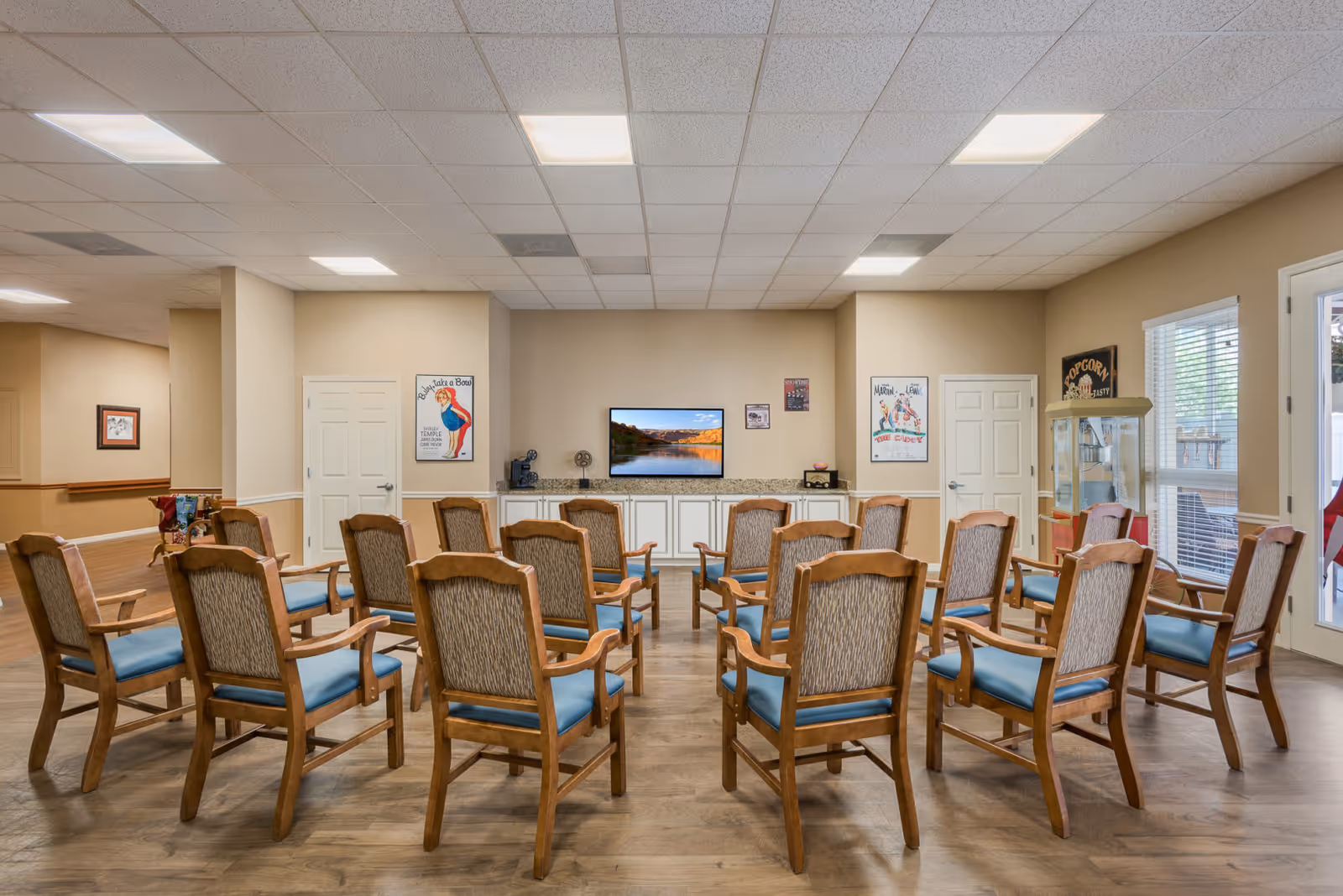 A common area with multiple wooden chairs arranged in rows facing a wall-mounted flat screen TV. The room has beige walls, a popcorn machine near a window, and vintage-style posters on the walls. The floor is wooden, and the ceiling has recessed lighting.
