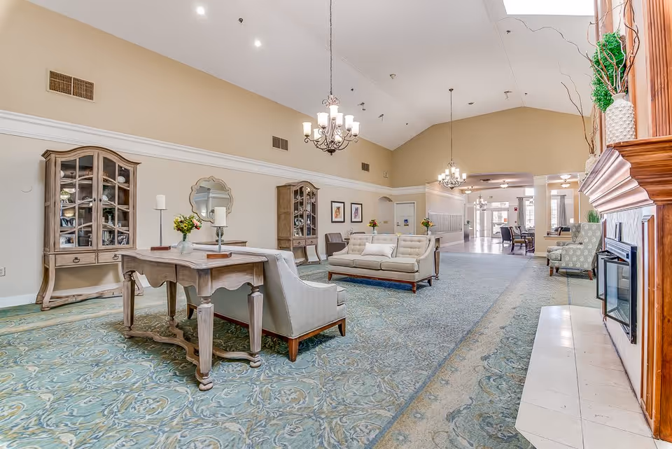 A spacious and well-lit senior living facility common area with high vaulted ceilings, chandeliers, and a large patterned carpet. The room features comfortable seating including sofas and armchairs, wooden display cabinets, a wooden table with candles and flowers, and a fireplace with decorative plants on the mantel. The area extends into a hallway with additional seating and natural light coming from the entrance.