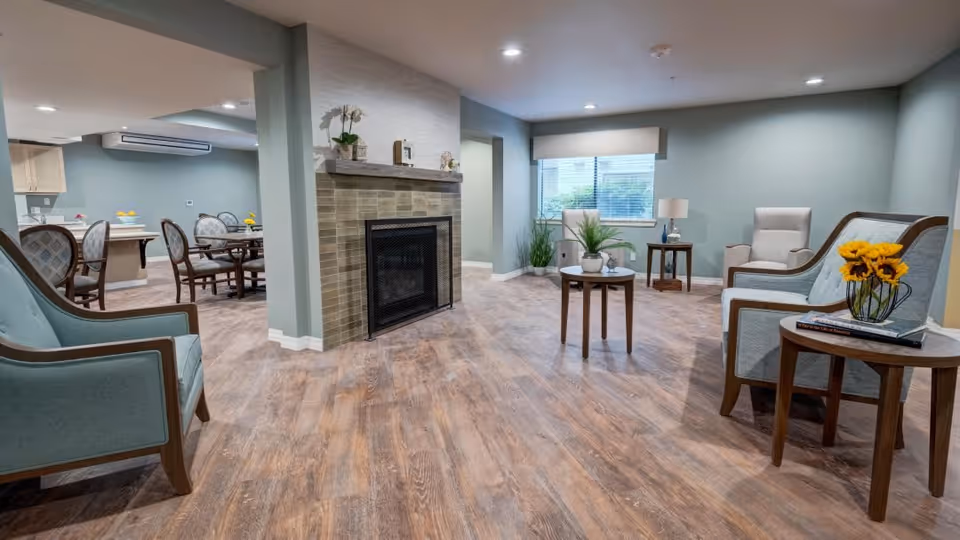 A spacious and well-lit common area with wooden flooring, light blue walls, and recessed ceiling lights. The room features a fireplace with decorative items on the mantel, several armchairs with wooden frames and light blue upholstery, small round tables with plants and flowers, and a window with blinds letting in natural light. In the background, there is a dining area with a table and chairs.