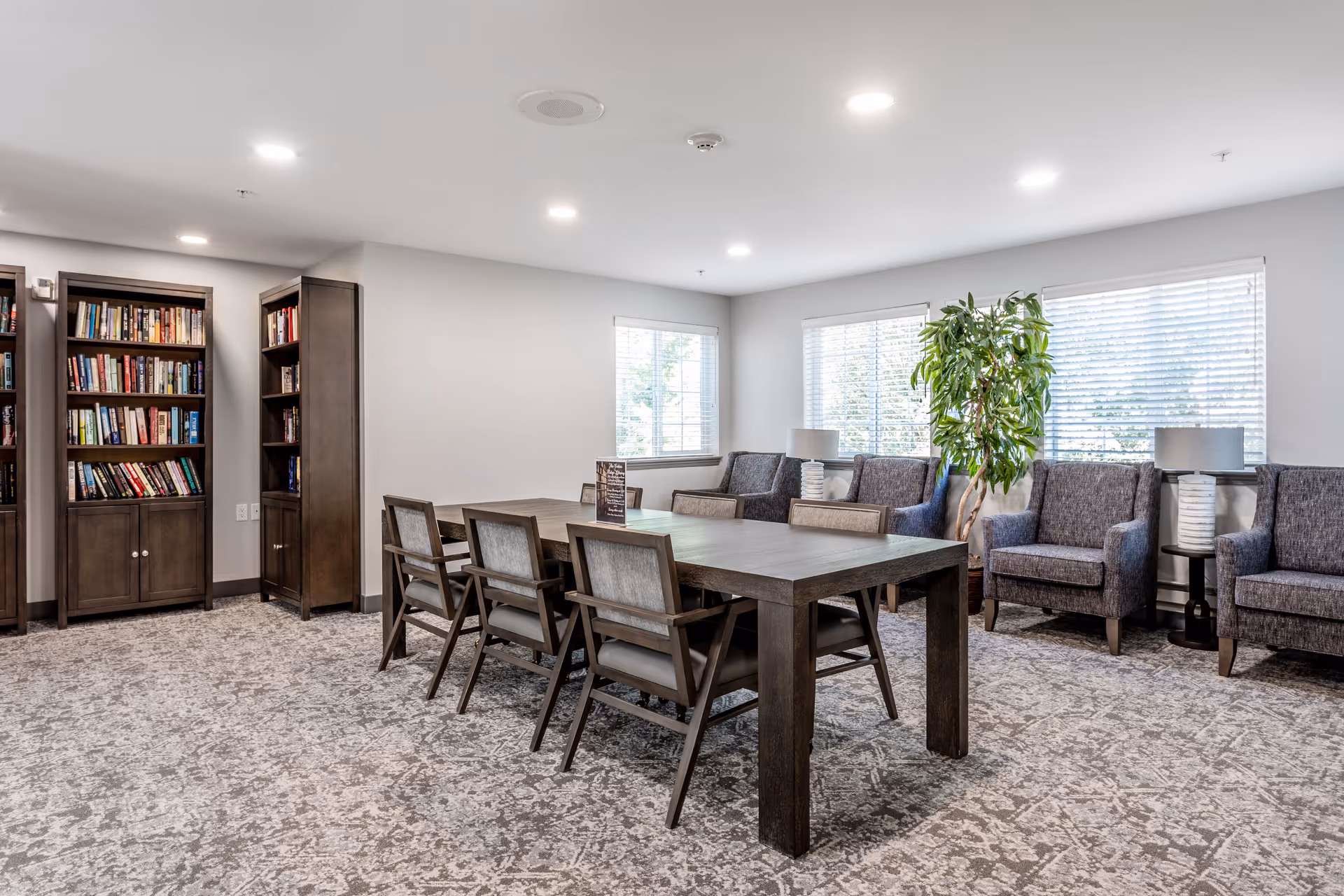 Bright communal lounge with a long wooden table, chairs, bookshelves, and upholstered armchairs by windows.