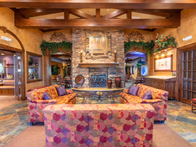 A cozy senior living facility common area with a stone fireplace as the focal point. The room features wooden beams on the ceiling, two patterned sofas with purple cushions facing each other, and a glass coffee table in the center. The walls are decorated with greenery and artwork, and there are wooden doors and windows leading to adjacent rooms.