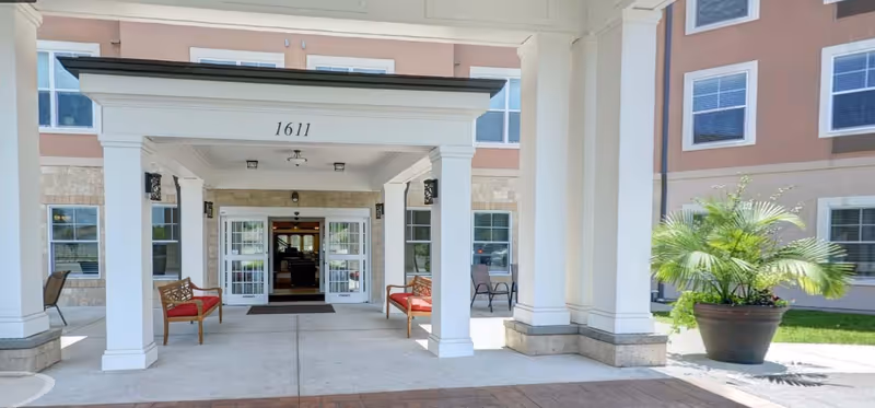 Entrance of a senior living facility with white pillars supporting a covered walkway. The building has large windows and beige walls with stone accents. Two wooden benches with red cushions are placed on either side of the entrance, and a large potted plant is visible on the right side. The number 1611 is displayed above the entrance.