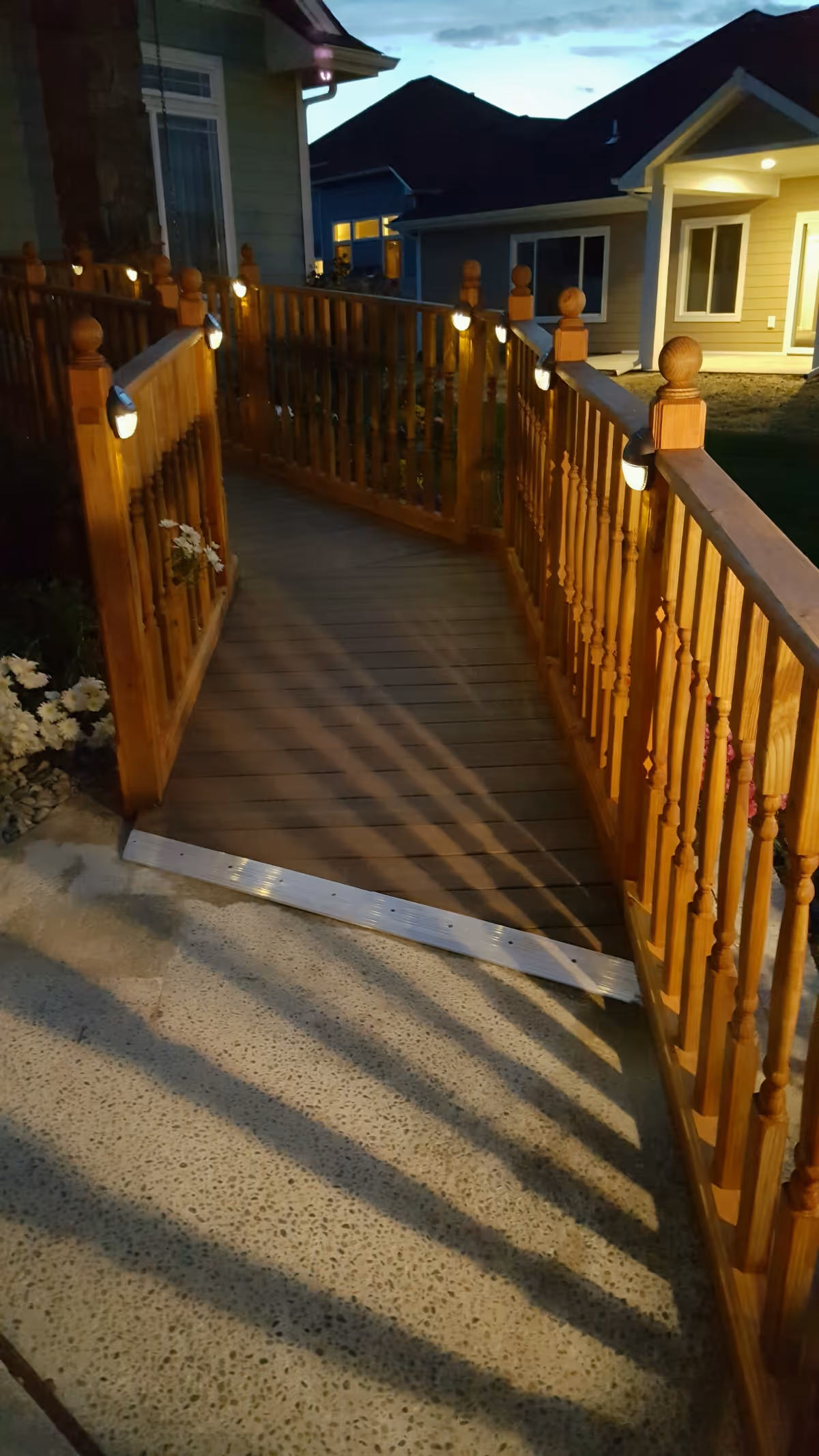 A wooden ramp with handrails on both sides, illuminated by small lights attached to the railings, leading up to the entrance of a building during dusk.