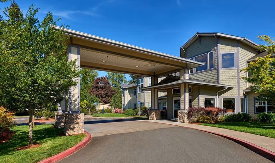 Exterior view of a senior living facility building with a covered entrance driveway, surrounded by green trees and landscaping under a clear blue sky.