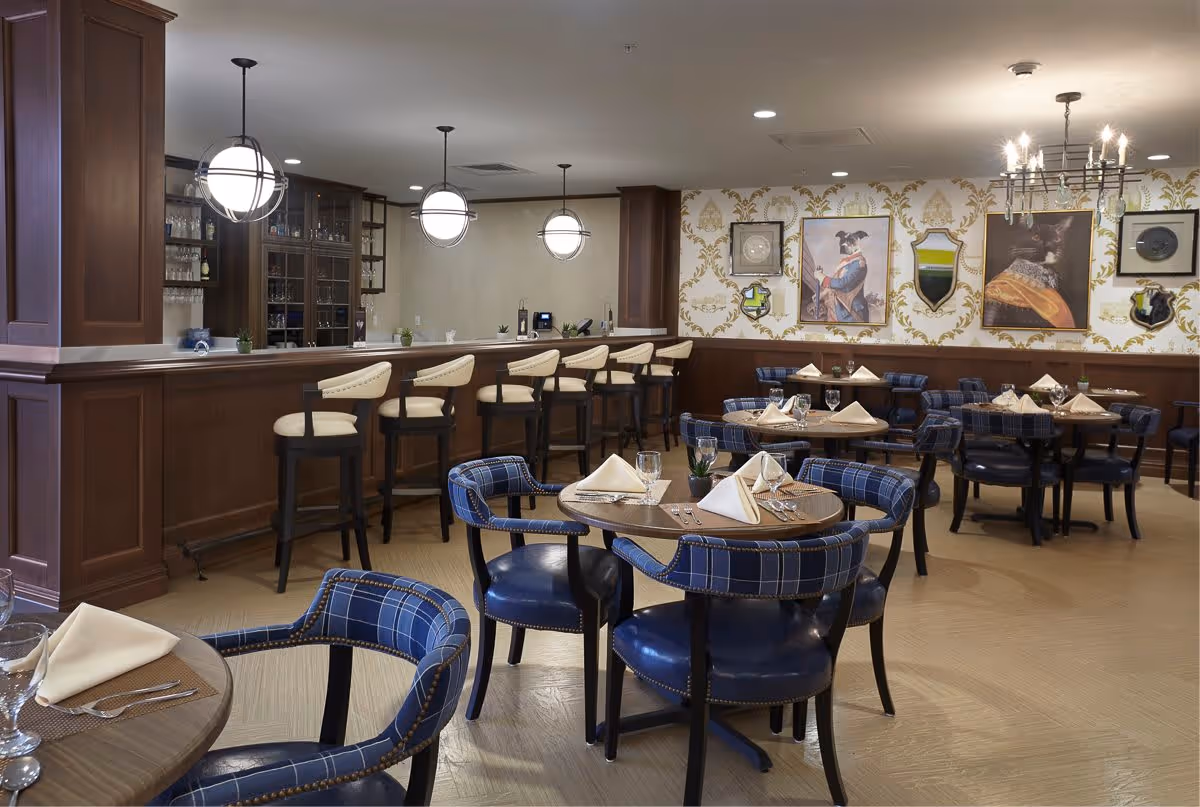 A dining room with round wooden tables set with folded napkins, glasses, and silverware. Blue plaid upholstered chairs surround the tables. There is a wooden bar with cream-colored cushioned bar stools and three spherical pendant lights hanging above. The walls have decorative wallpaper with framed artwork and mirrors.