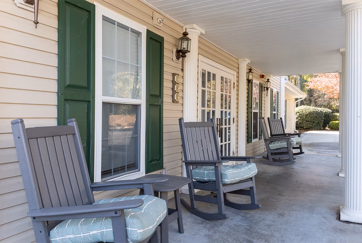A covered porch area with several gray rocking chairs featuring light blue patterned cushions. The porch has white columns, beige siding with green shutters, and multiple windows and glass doors. There are outdoor wall lanterns mounted on the wall.