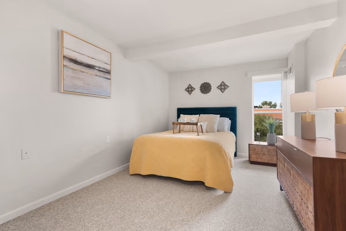 A cozy bedroom featuring a yellow quilted bedspread, a teal headboard, and decorative wall art. A bedside table holds a tray with items, and a window provides natural light, showcasing greenery outside.