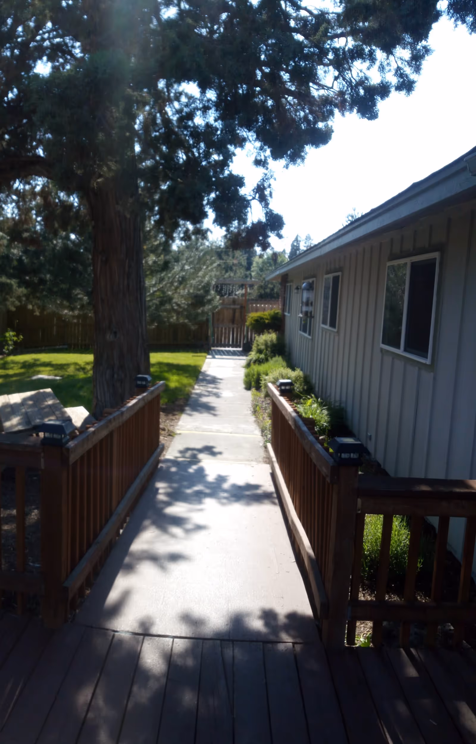 A sunny outdoor pathway with a wooden railing on both sides leading to a sidewalk. To the right is a beige building with several windows, and to the left is a large tree and a grassy area with a wooden fence in the background.