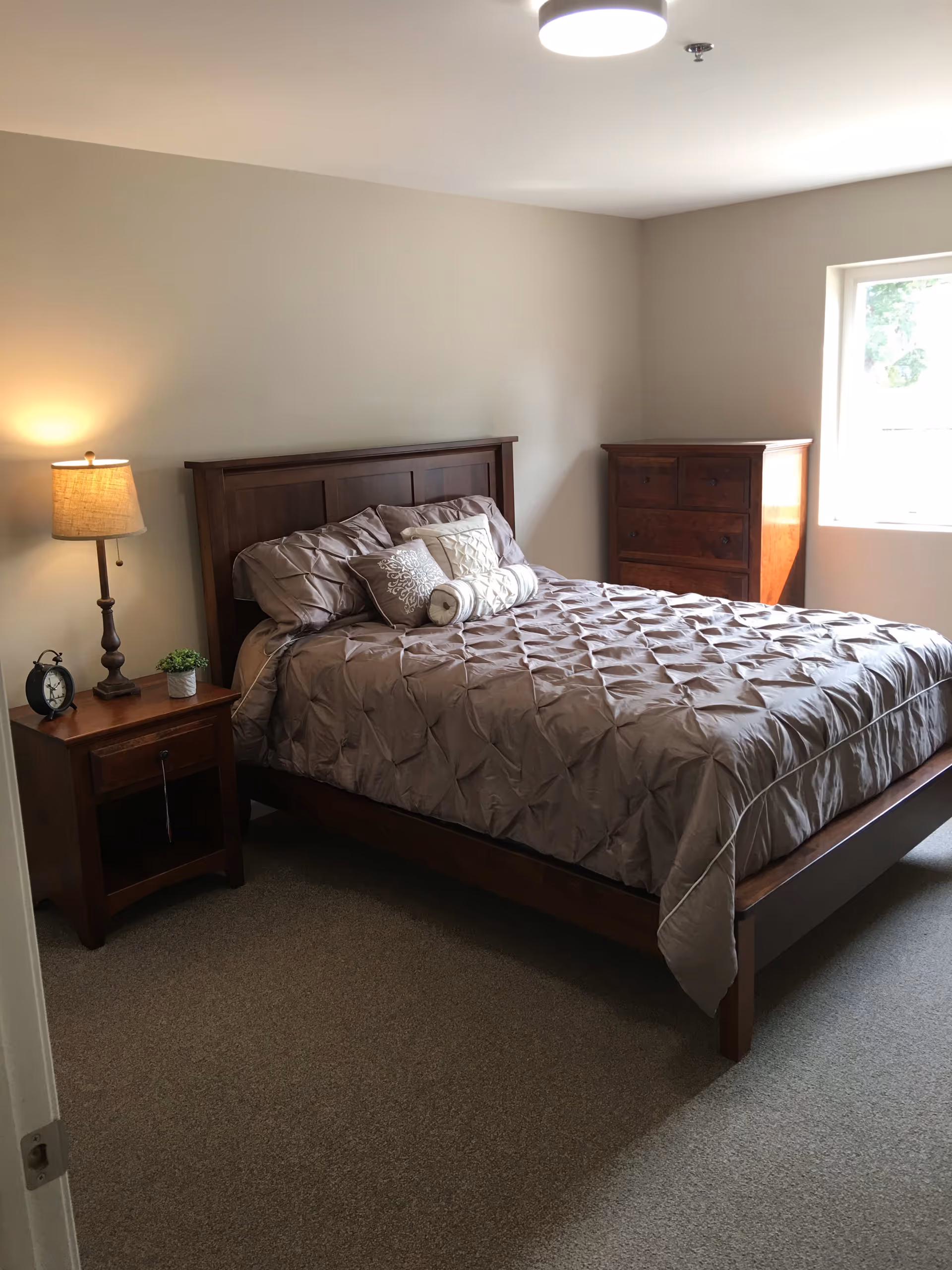 A bedroom with a wooden bed frame and a neatly made bed with beige bedding and decorative pillows. To the left of the bed is a wooden nightstand with a lamp, a small plant, and an alarm clock. On the right side of the room is a wooden dresser next to a window letting in natural light. The walls are painted light beige and the floor is carpeted.