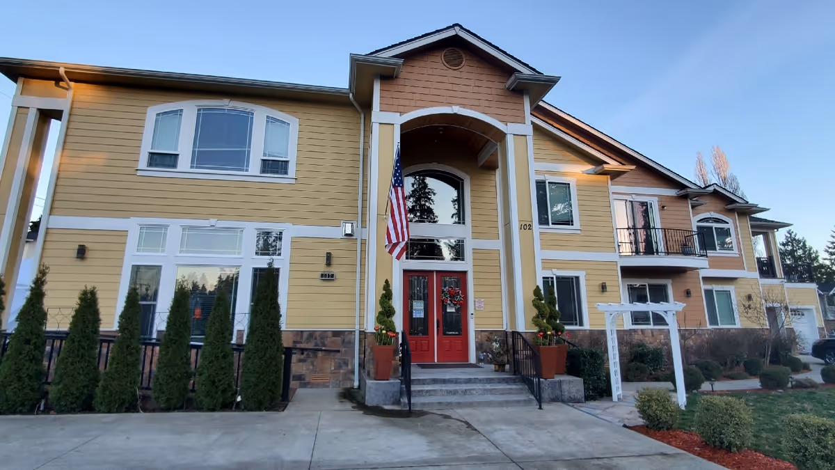 Exterior front view of a two-story senior living facility building with yellow and brown siding, white trim, and a red double-door entrance. There are potted plants and an American flag near the entrance, with a concrete walkway and landscaped bushes in front.