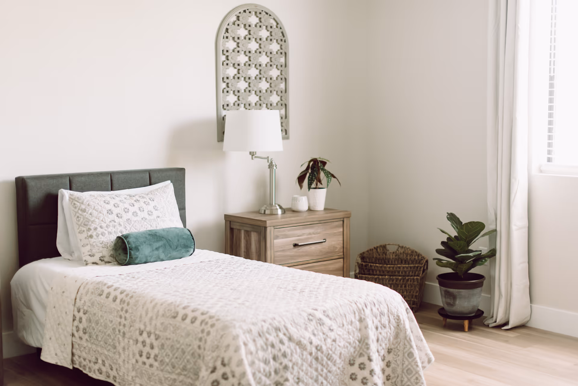 Sunlit single bedroom with a neatly made bed, wooden nightstand with a lamp and plants, a woven basket and potted plant by a window.