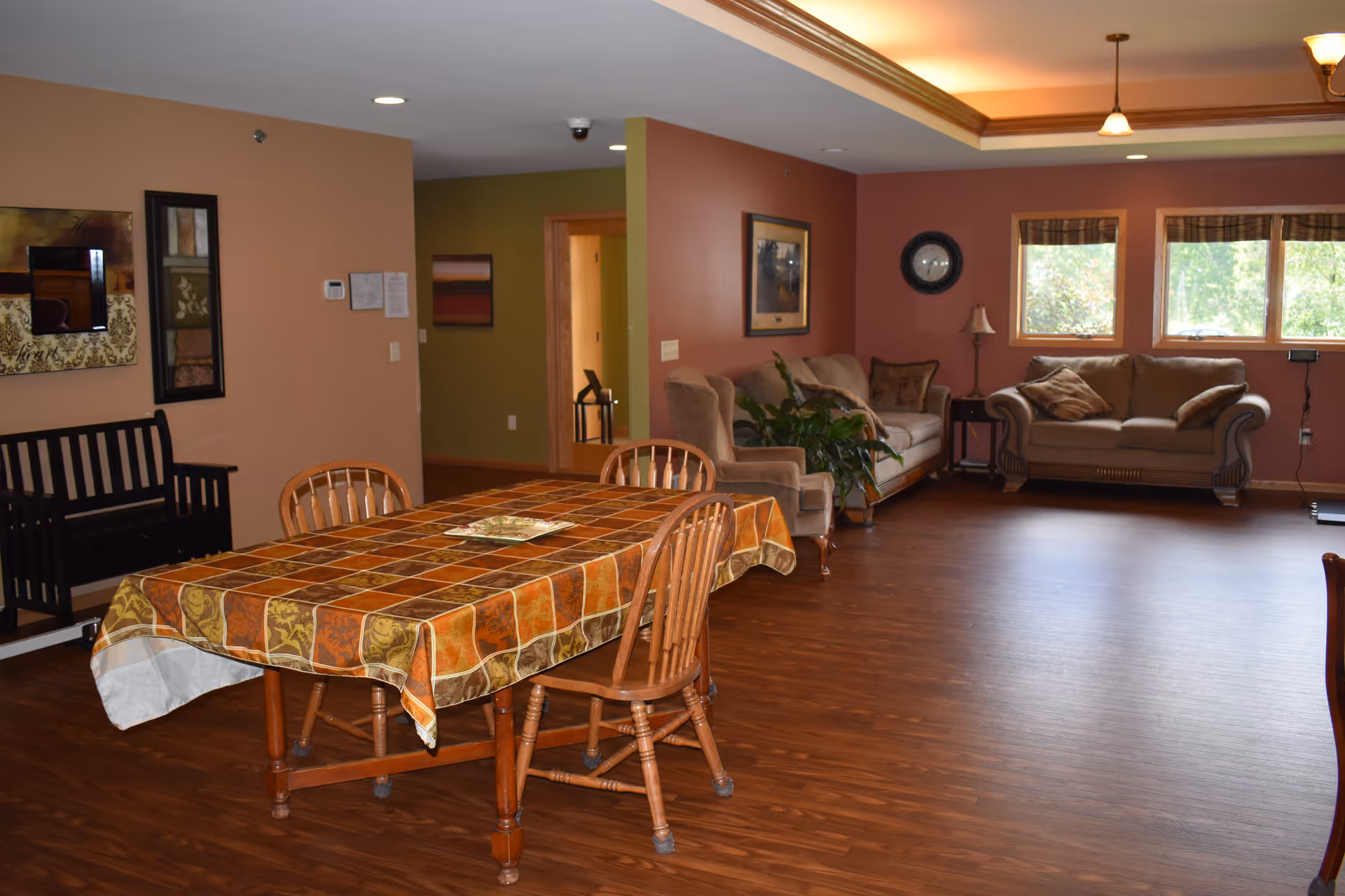 A spacious room with a wooden floor featuring a dining table covered with a patterned tablecloth and surrounded by wooden chairs. In the background, there are two sofas with cushions, a small side table with a lamp, a wall clock, and three windows letting in natural light. The walls are painted in warm tones of brown and green, and there is framed artwork on the walls.