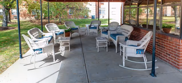 Outdoor covered patio area with white wicker chairs and small tables arranged in a semi-circle on a concrete floor, surrounded by grass and trees with a brick building in the background.