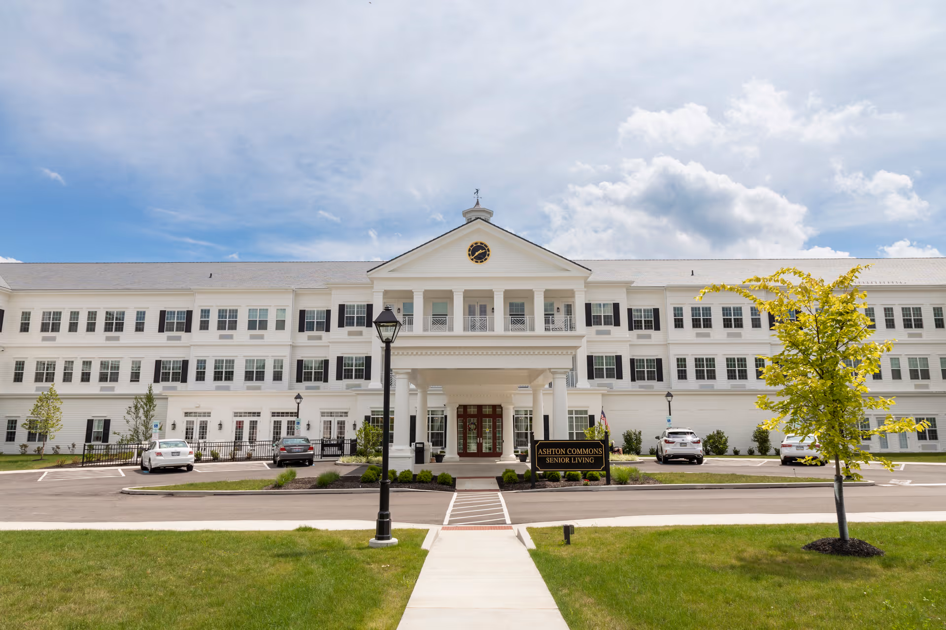 Front entrance of a white multi-story senior living building with columns, a driveway, parked cars, and a sign for Ashton Commons.