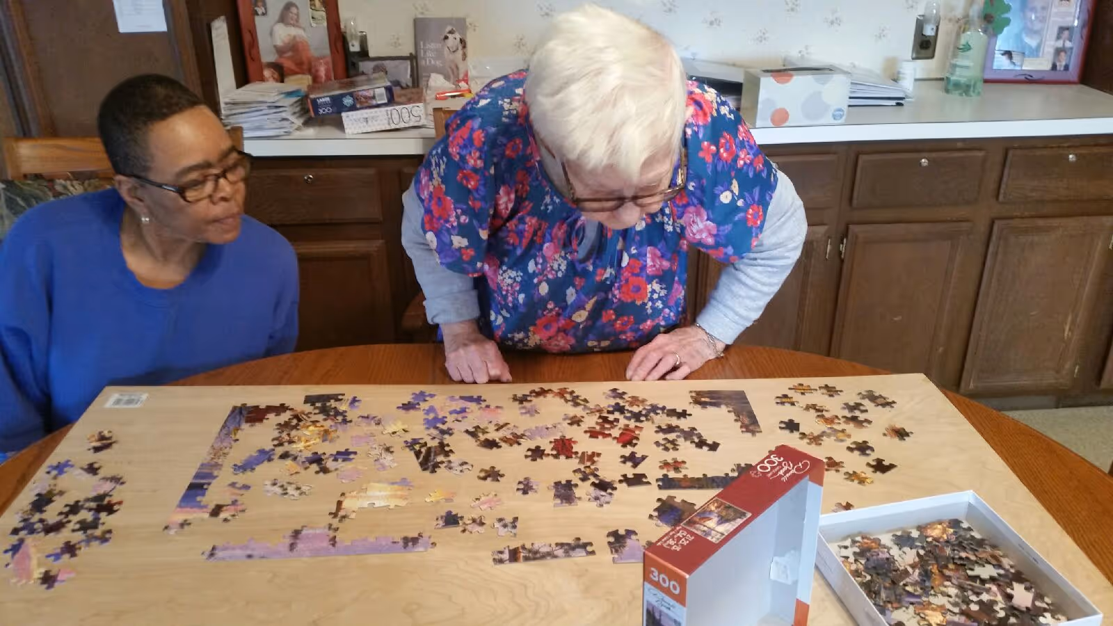 Two elderly women working together on assembling a 300-piece jigsaw puzzle on a wooden table in a room with wooden cabinets and various items on the countertop in the background.