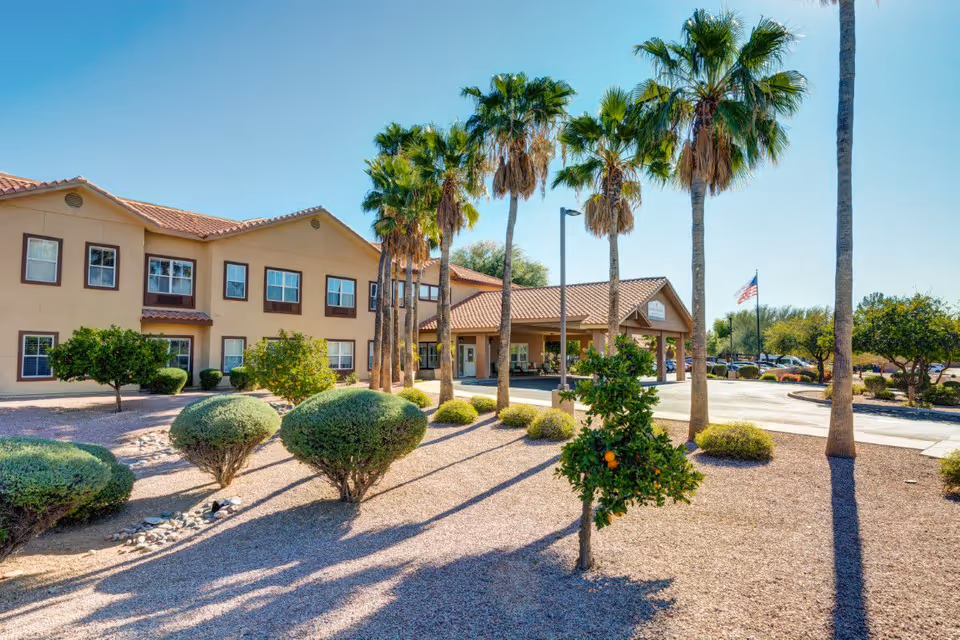 Exterior view of Pegasus Landing of Mesa facility showing a two-story building with beige walls and brown trim, surrounded by palm trees, bushes, and desert landscaping under a clear blue sky.