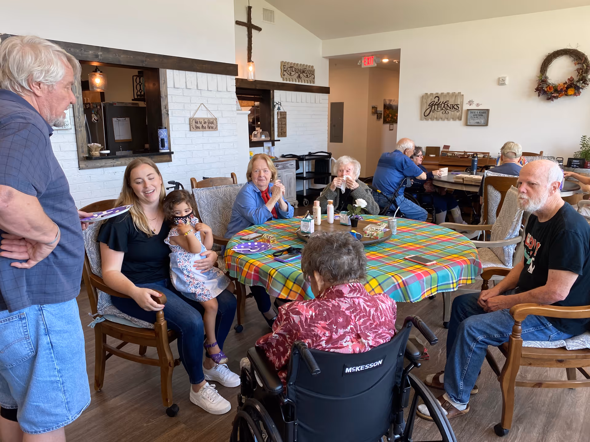 A group of elderly people and a young woman with a child gathered around a table covered with a colorful plaid tablecloth in a communal dining area. The room has white walls decorated with signs and a wreath, and there is a cross mounted above a pass-through window to the kitchen. Some people are seated in chairs, one person is in a wheelchair, and others are standing or sitting at another table in the background.