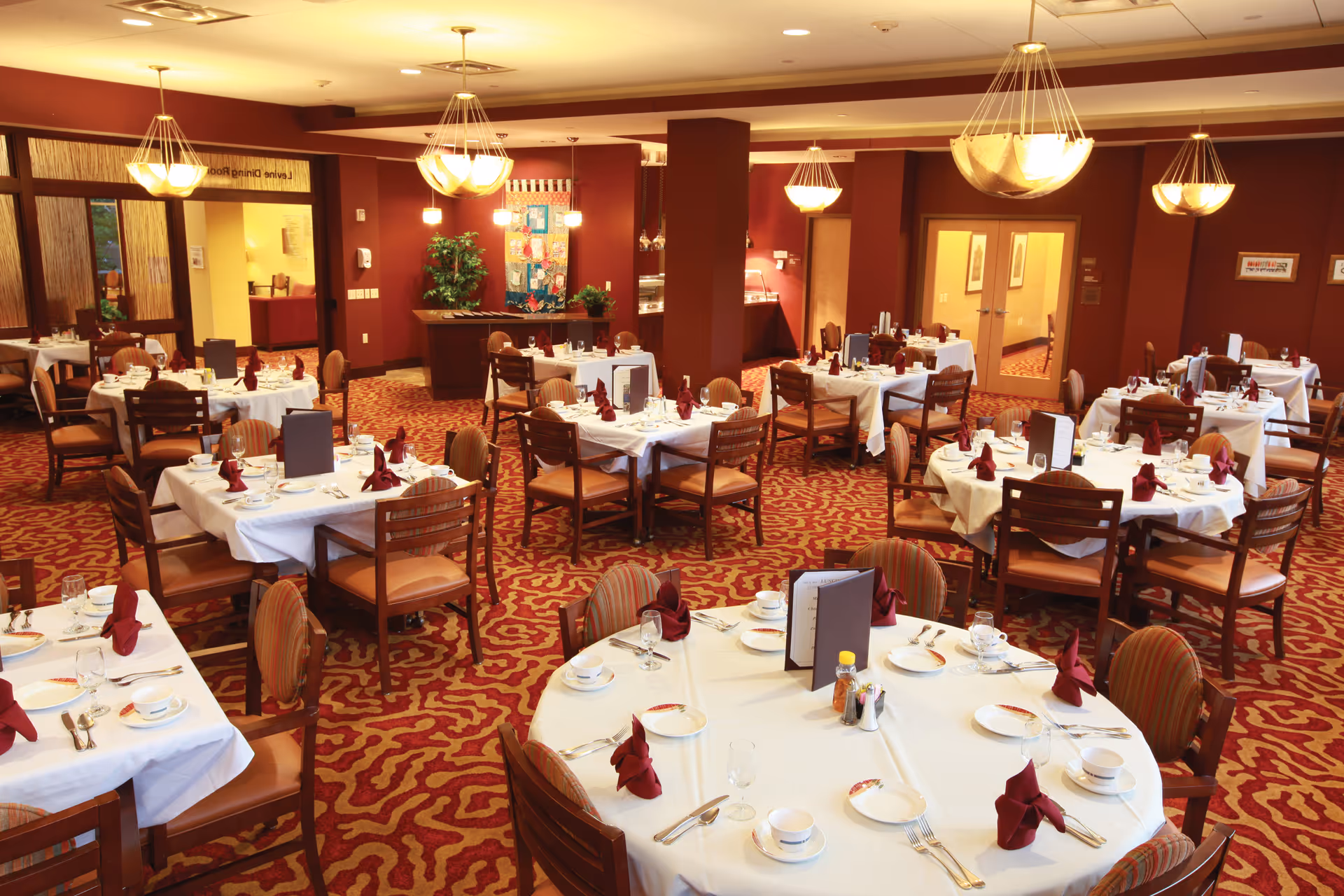 A spacious dining room with multiple round and square tables covered with white tablecloths, each set with plates, cups, silverware, and folded maroon napkins. The room has warm lighting from hanging chandeliers and a patterned red and gold carpet. There are wooden chairs with cushioned seats around the tables, and a serving area is visible in the background.