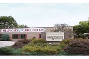 Exterior view of Shady Oaks Care Center building with a sign in front surrounded by bushes and greenery under a clear sky.