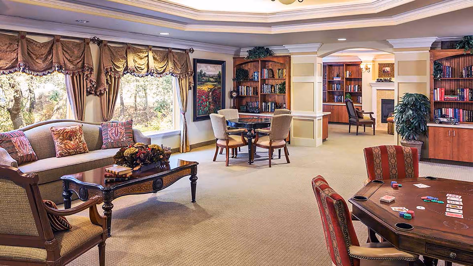 Sunlit communal lounge with sofas, armchairs, bookshelves, and a card table set up for a game.