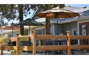 Outdoor patio area with wooden fencing, tables, chairs, and large umbrellas providing shade in front of a building with multiple windows and doors.
