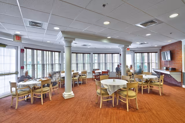 A spacious dining room with round tables covered with white tablecloths and wooden chairs. Large windows with vertical blinds allow natural light to fill the room. Several elderly people are seated at the tables, engaged in conversation. The room features white columns, a red carpet, and a wall-mounted television on a brick accent wall.