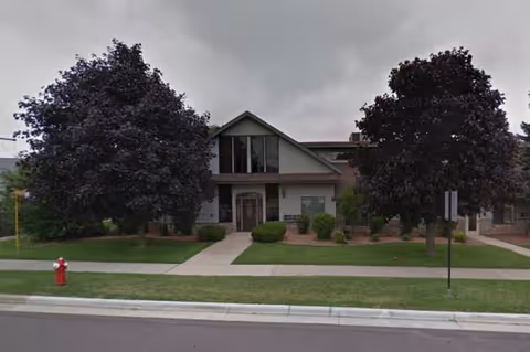 Front view of a two-story facility with peaked roof, trees, and a sidewalk.