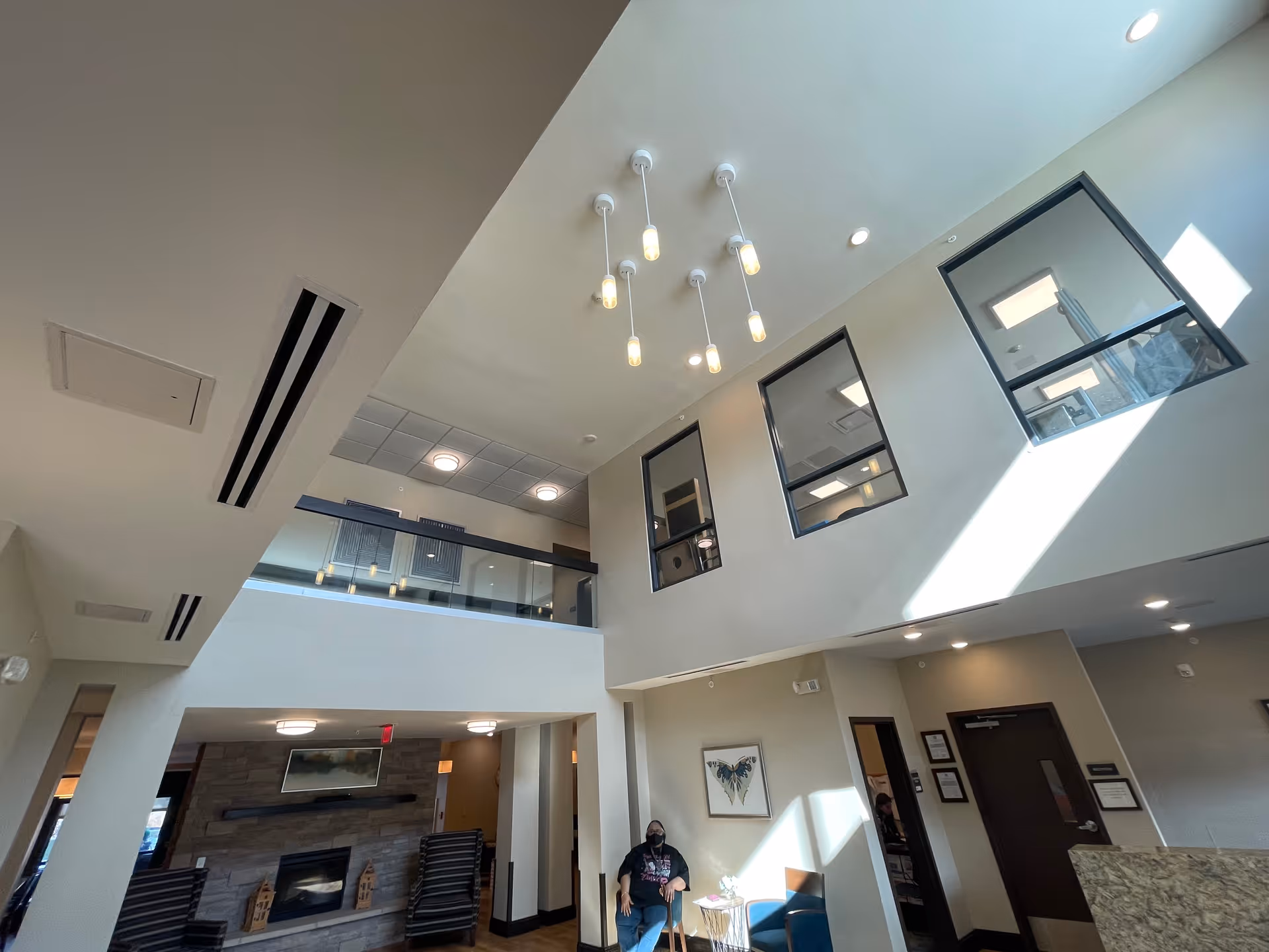 Interior view of a senior living facility lobby with high ceilings and modern hanging lights. There is a fireplace with chairs arranged around it, a person sitting on a chair near a small table, and windows on the upper level letting in natural light.