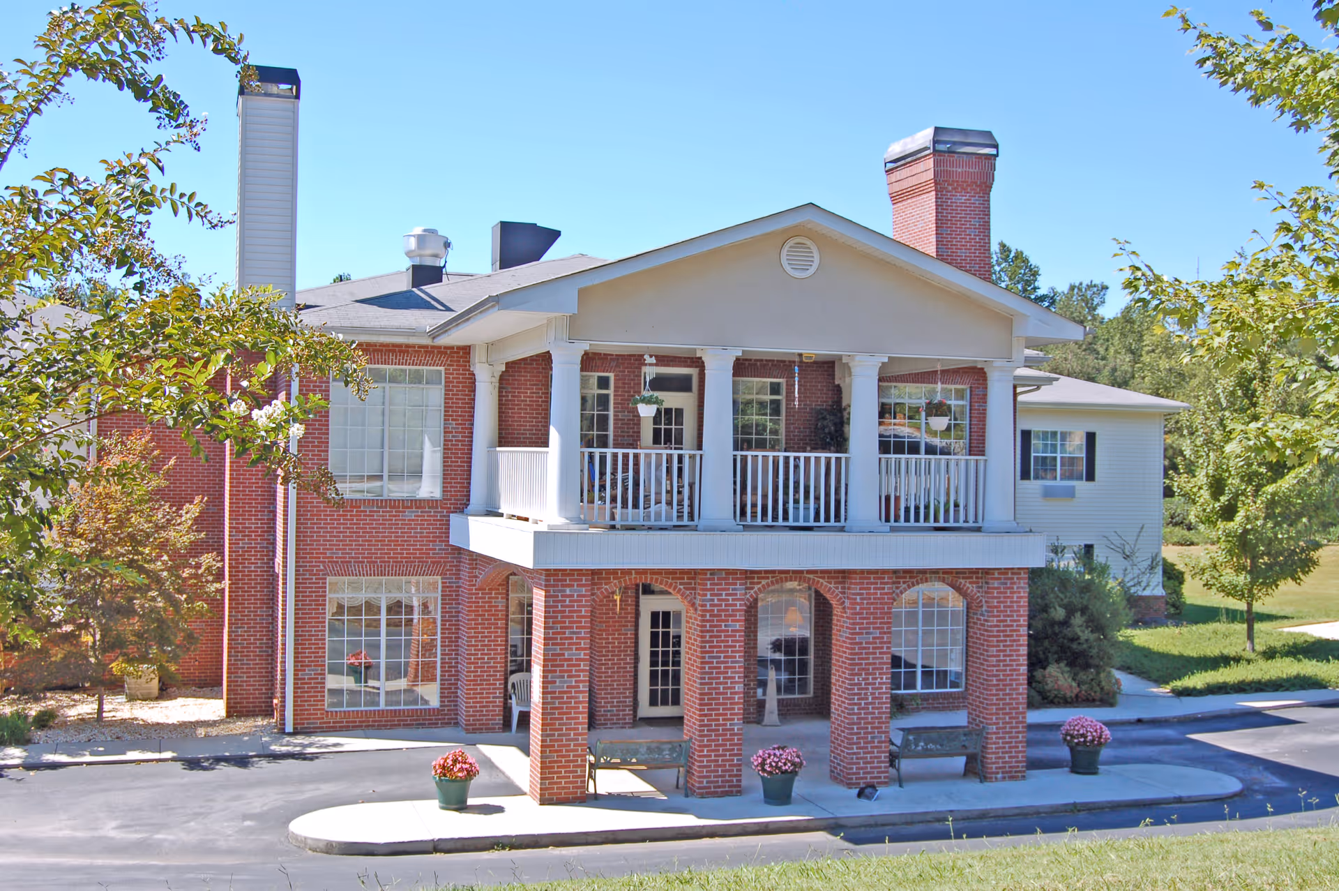 Two-story red brick assisted living building with a covered balcony and columned entrance flanked by potted flowers.