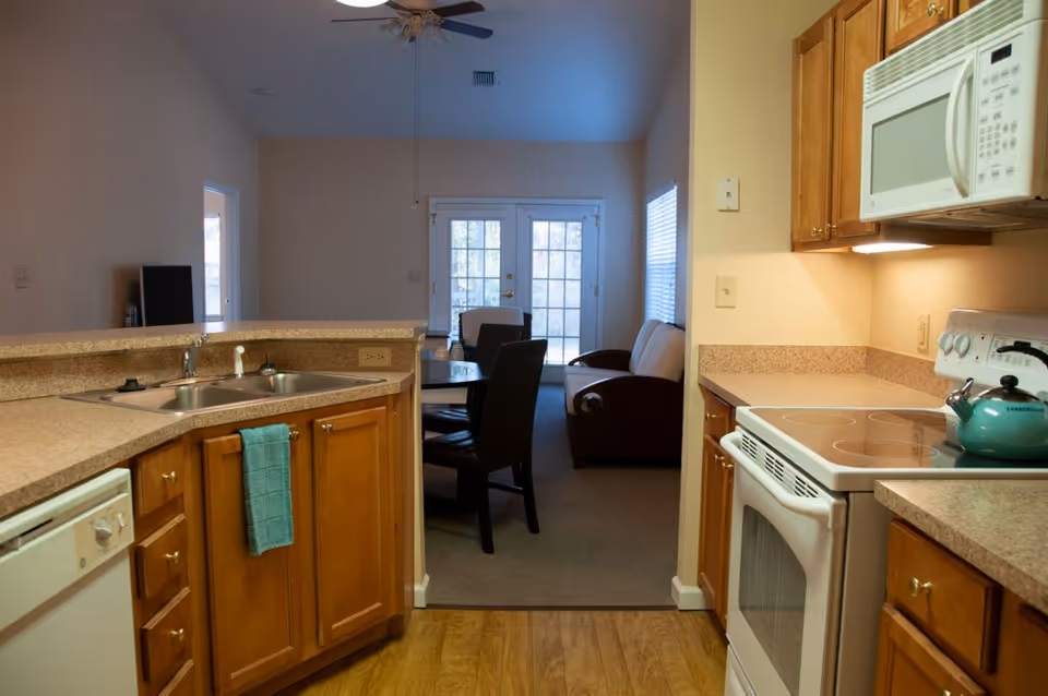 View of a kitchen with wooden cabinets, a white stove with a teal kettle, a microwave, and a double sink with a blue towel hanging on the cabinet. Beyond the kitchen is a dining area with a round table and chairs, and a living room with a sofa and armchair near glass doors leading outside.