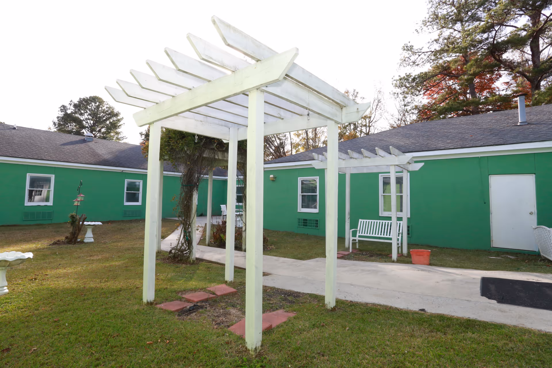 Outdoor courtyard area of St. Francis Assisted Living Community featuring a white wooden pergola, green building walls with white-trimmed windows and doors, a white bench, bird feeders, and a birdbath on a grassy lawn with a concrete walkway.