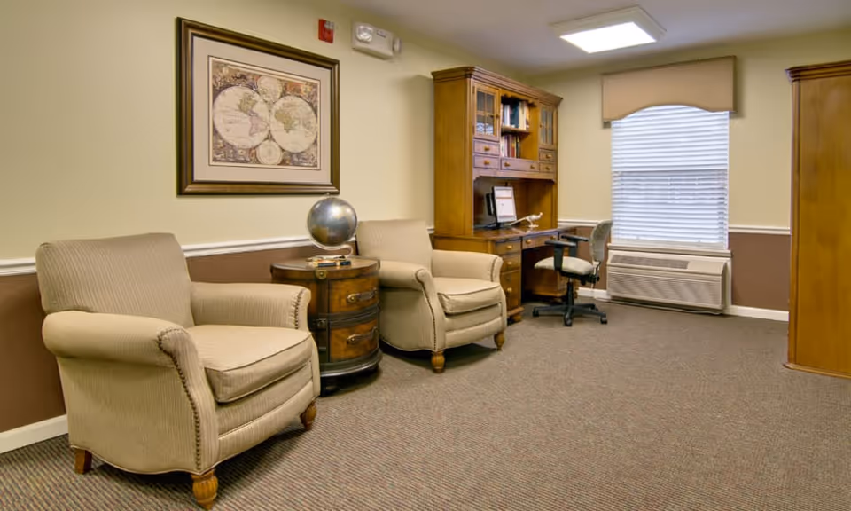 Seating area with two beige armchairs flanking a round side table topped with a globe, and a wooden desk and office chair by a window.