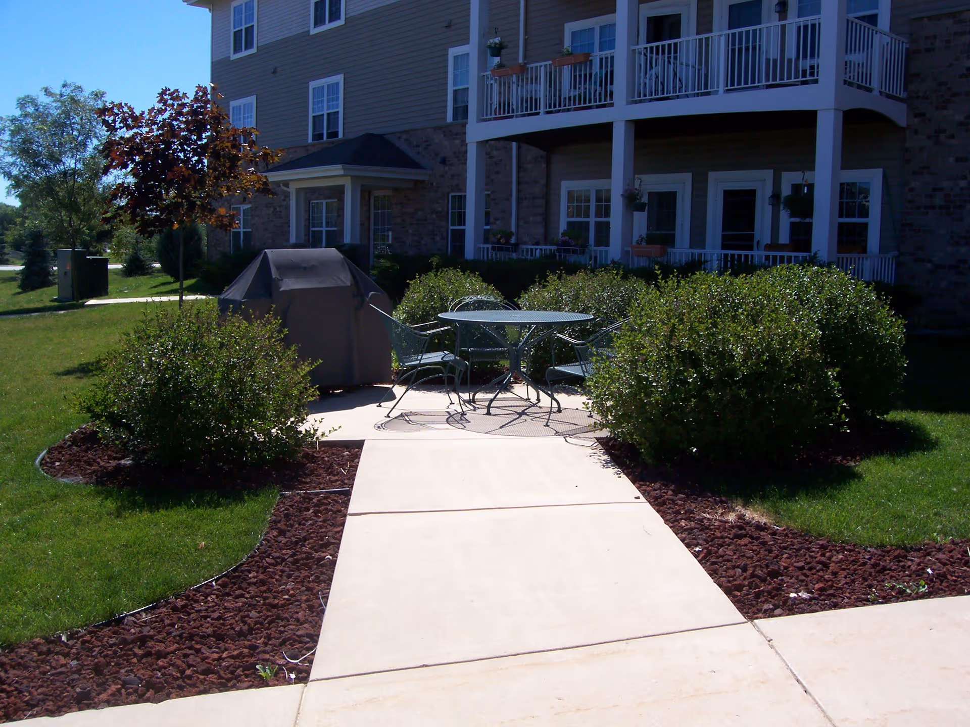 Outdoor patio area with a metal table and four chairs surrounded by bushes and a covered grill. The patio is adjacent to a multi-story residential building with balconies and windows. The area is landscaped with grass, trees, and mulch beds.