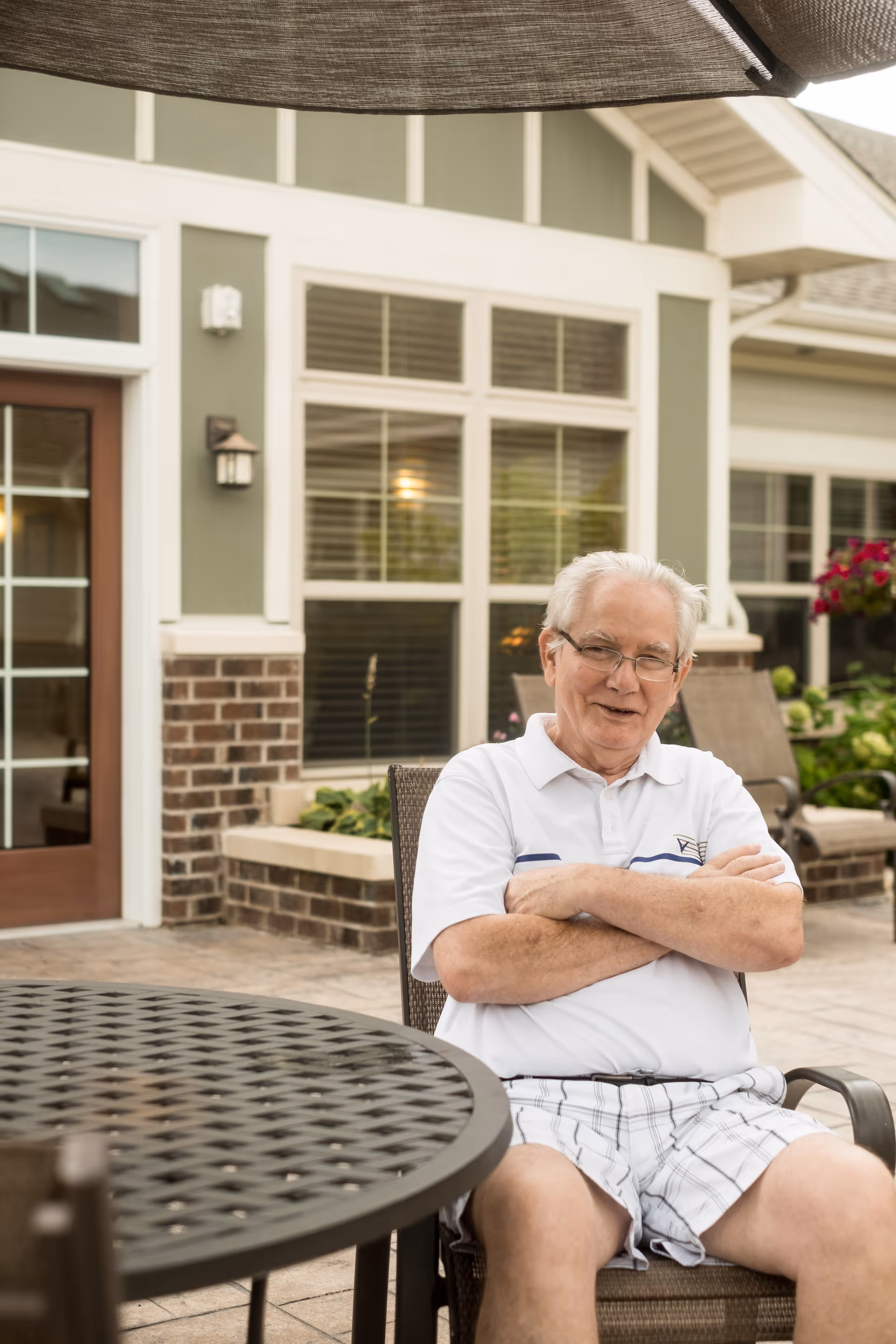 An elderly man with white hair and glasses sits outdoors on a patio chair with his arms crossed, smiling. He is wearing a white polo shirt and plaid shorts. Behind him is a building with large windows, brick and green siding, and a door with glass panels. There are plants and flowers near the building and a round metal patio table in the foreground.