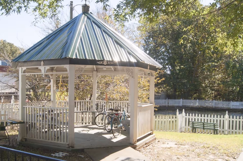 A white wooden gazebo with a green metal roof situated outdoors on a sunny day. Inside the gazebo, there are two bicycles parked. Surrounding the gazebo is a white picket fence, green grass, trees, and a green bench near the fence.