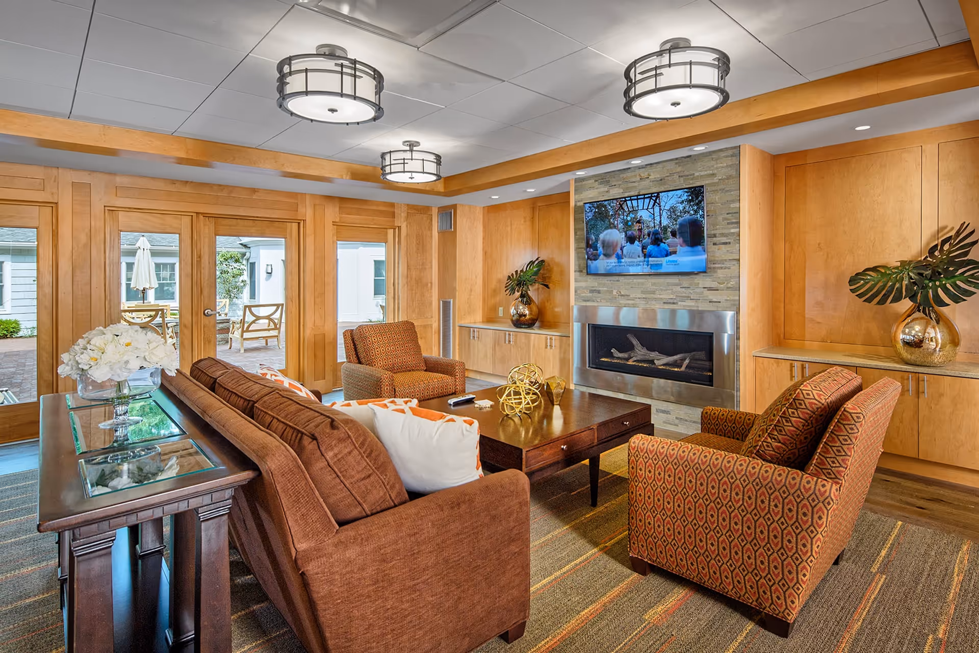A cozy living room with a brown sofa and two patterned armchairs arranged around a wooden coffee table. A flat-screen TV is mounted above a modern fireplace on a stone accent wall. The room features wooden paneling, built-in cabinets with decorative vases, and large glass doors leading to an outdoor patio area.
