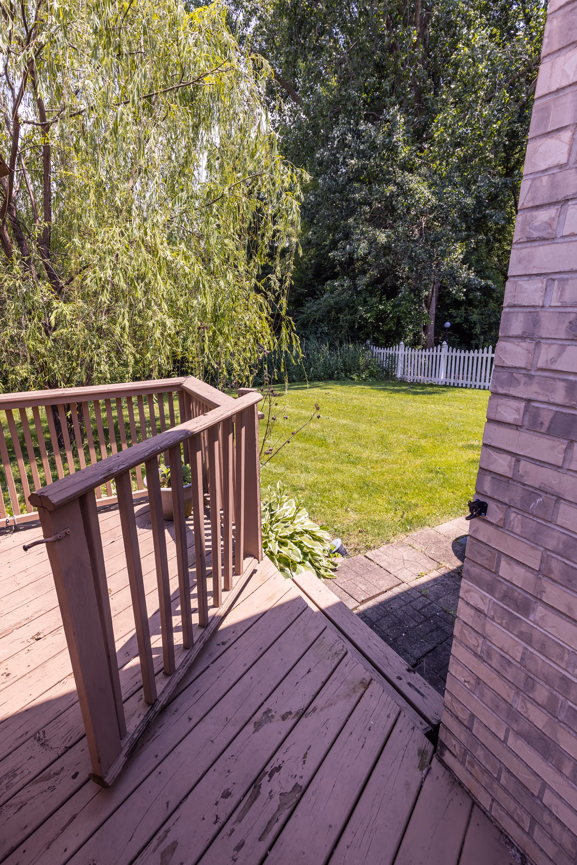 View from a wooden deck with railings looking out onto a green lawn bordered by trees and a white picket fence. The deck is attached to a brick building, and there is a small paved pathway leading from the deck to the lawn.