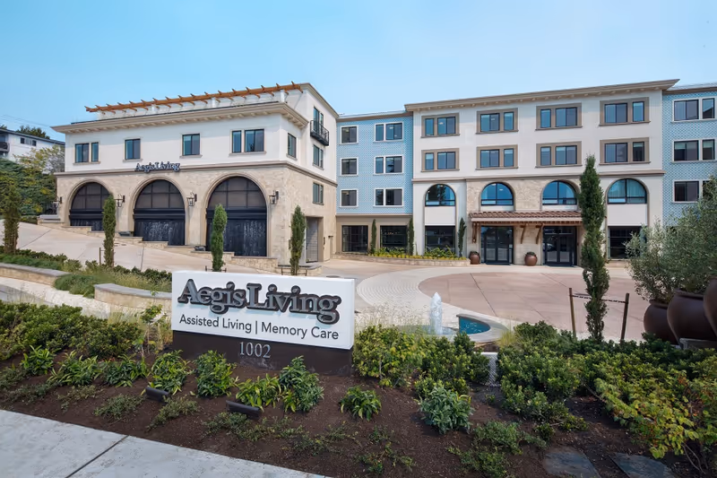 Exterior view of Aegis Living Kirkland Waterfront, a multi-story assisted living and memory care facility with landscaped greenery, a water fountain, and a prominent sign displaying the facility name and address.
