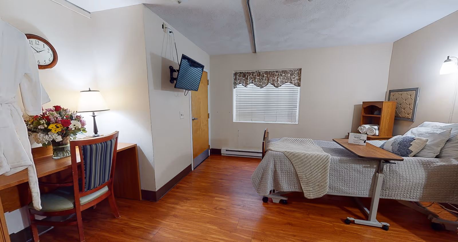 A senior living facility bedroom with a hospital-style bed covered in a gray quilt and pillows. There is a wooden overbed table with rolled towels and a sign on it. A wooden chair with a striped cushion is placed at a desk with a vase of flowers and a lamp. A wall-mounted TV is angled towards the bed. The room has wood flooring, a window with blinds and a valance, and a clock on the wall.