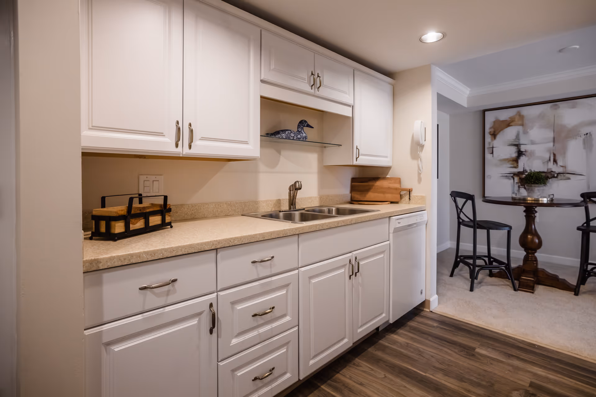 A kitchen area with white cabinets and drawers, a double sink, and a beige countertop. On the countertop are a wooden cutting board and a decorative item shaped like a duck. To the right, there is a small dining area with a round wooden table, two black chairs, and a large abstract painting on the wall. The floor is wood laminate in the kitchen and carpeted in the dining area.