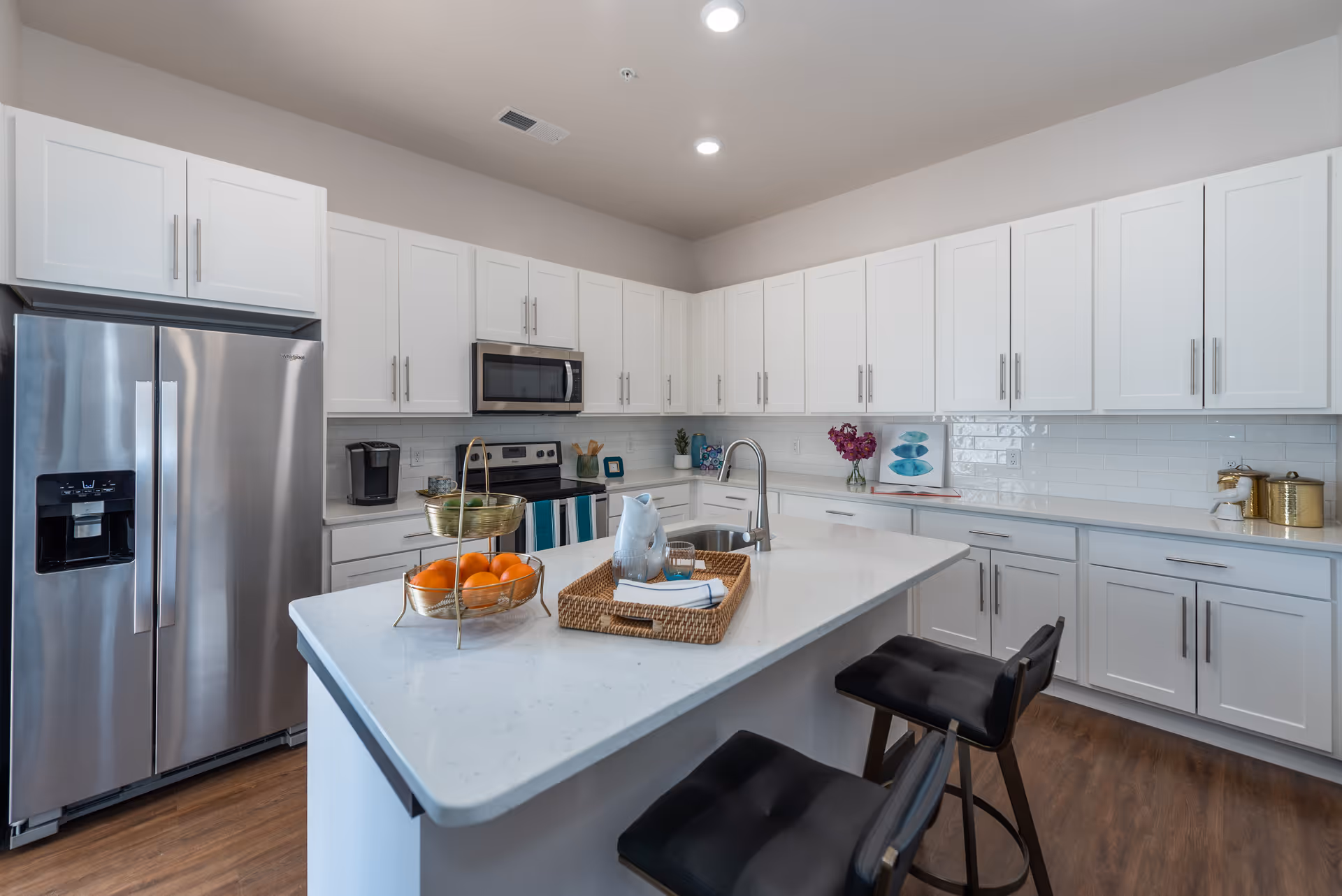 Modern kitchen with white cabinets, stainless steel refrigerator, microwave, and stove. A large white island countertop with a sink, two black cushioned bar stools, a wicker tray with a pitcher and glass, and a two-tiered fruit basket with oranges and limes. The kitchen has wood flooring and recessed ceiling lights.