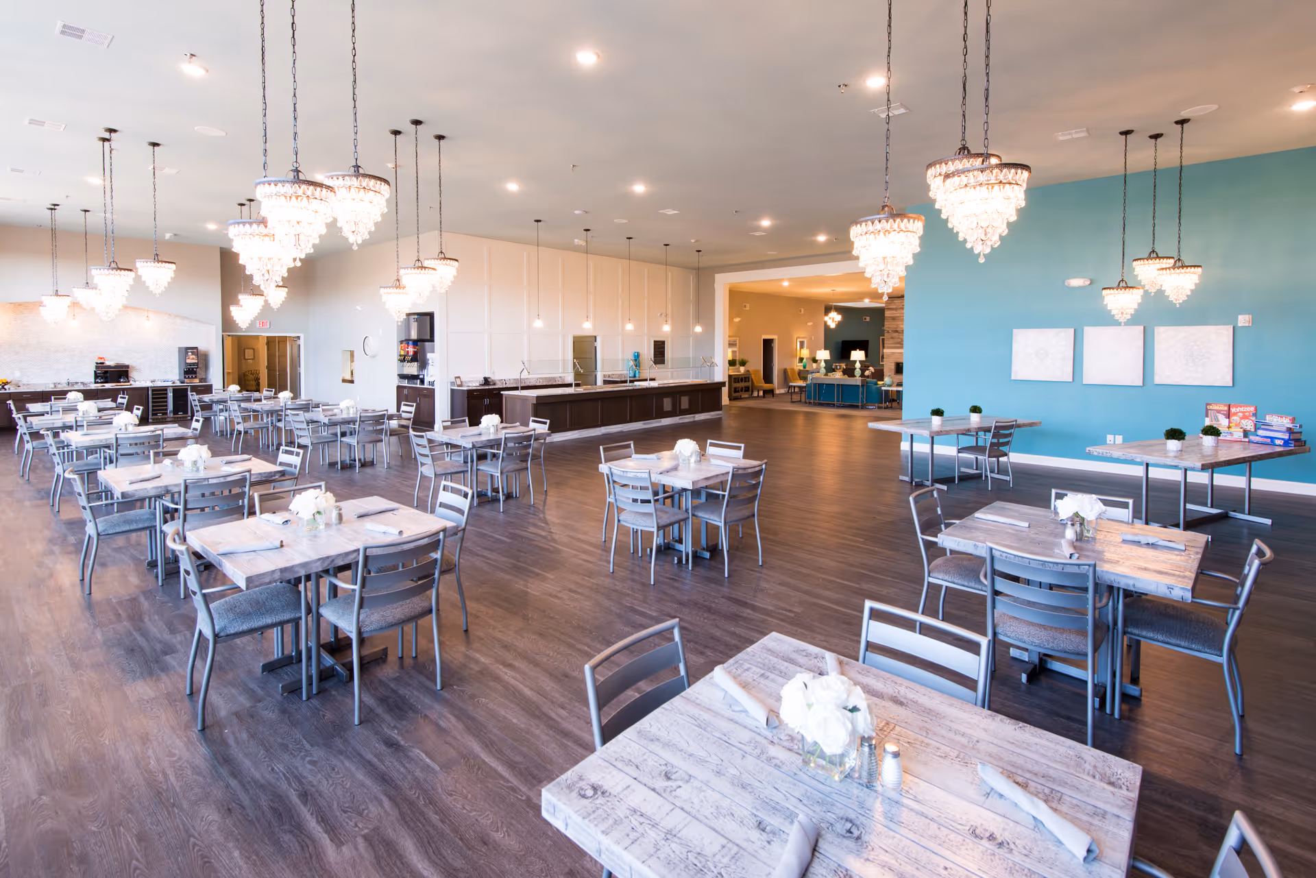 A spacious dining room with multiple tables and chairs arranged neatly. The tables have white floral centerpieces and rolled napkins. Elegant chandeliers hang from the ceiling, providing warm lighting. The room features a wooden floor, a teal accent wall with three framed artworks, and an open kitchen area in the background.
