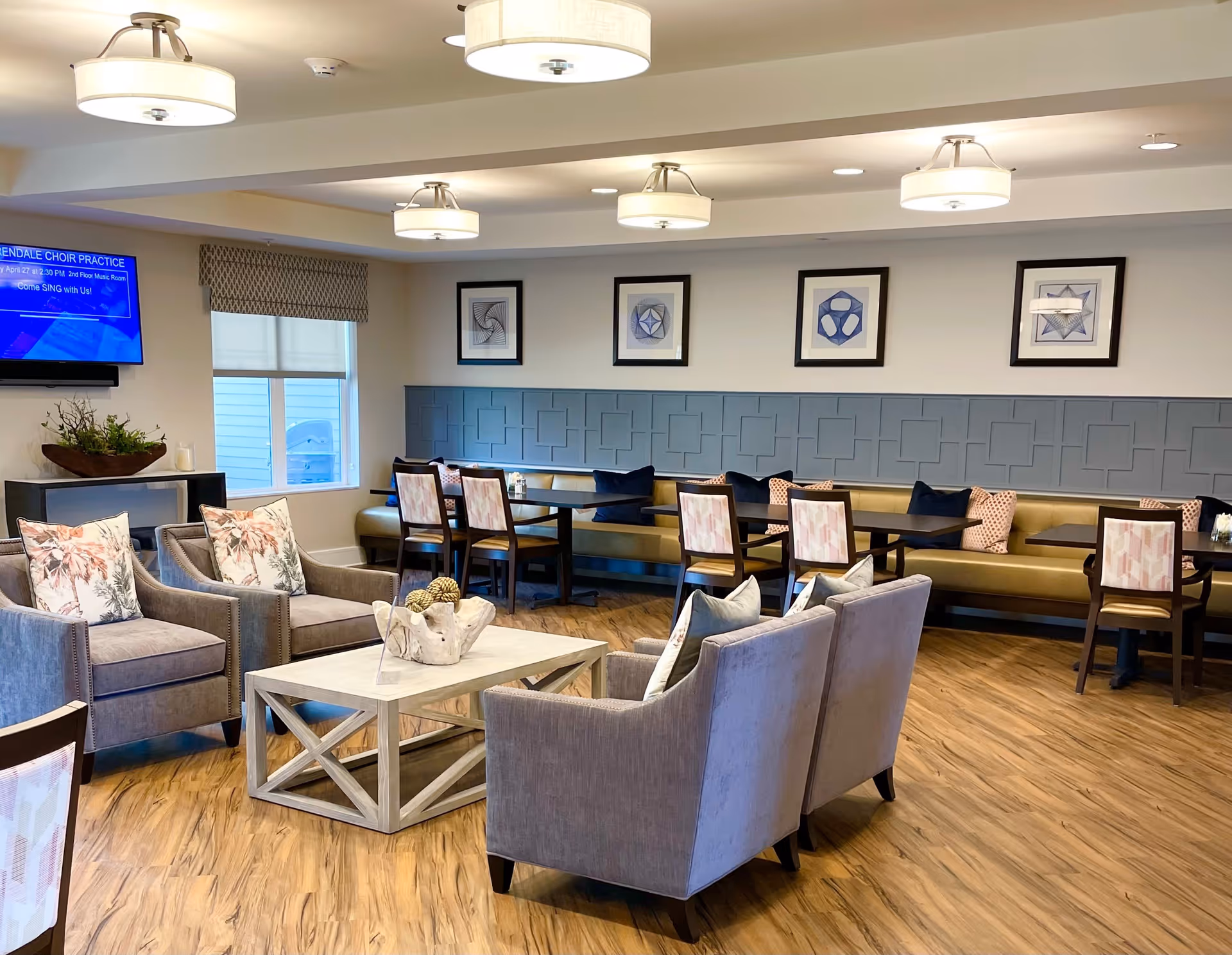 A well-lit common area with comfortable seating including armchairs and a sofa around a wooden coffee table. There are dining tables with chairs and a long bench along the wall, decorated with cushions. The walls have framed geometric artwork and a mounted TV screen displaying information about choir practice. The floor is wooden, and the ceiling has multiple modern light fixtures.