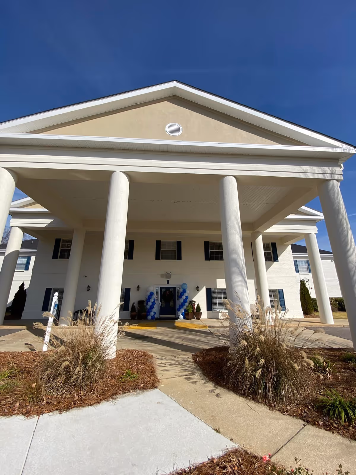 Front exterior view of a two-story building with large white columns supporting a covered entrance. The building has beige and white walls, black window shutters, and a clear blue sky above. There are decorative plants and bushes on either side of the walkway leading to the entrance, which is decorated with blue and white balloons.