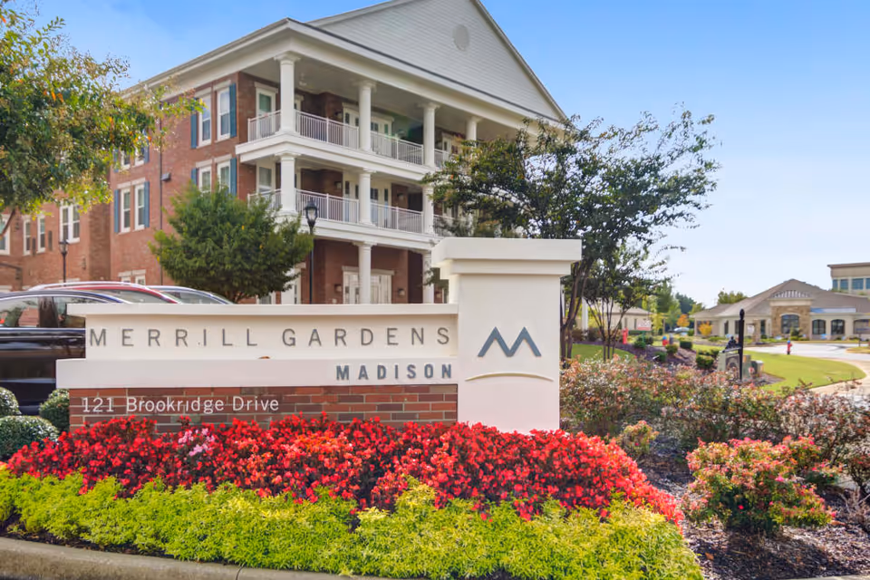 Exterior view of Merrill Gardens at Madison senior living facility showing a large brick and white columned building with balconies, a landscaped garden with red and green plants, and a sign that reads 'Merrill Gardens Madison 121 Brookridge Drive' under a clear blue sky.