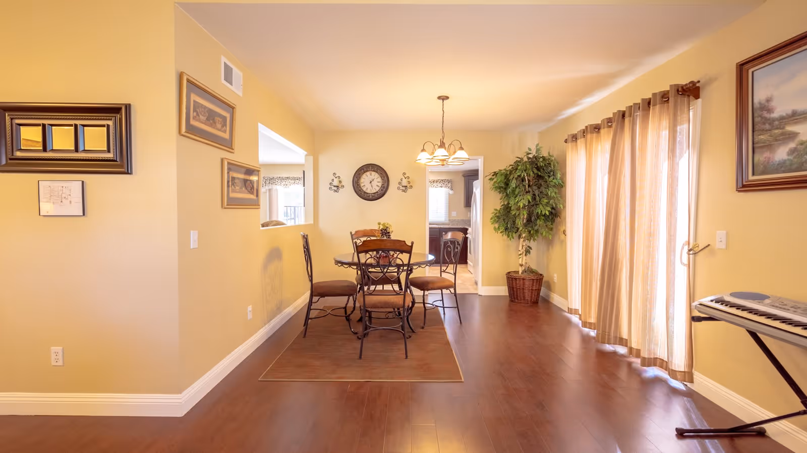 A cozy dining area with a round table and four chairs on a rug, beige walls decorated with framed pictures and a clock, a large potted plant in the corner, sheer curtains covering a sliding glass door, and a keyboard on a stand to the right.