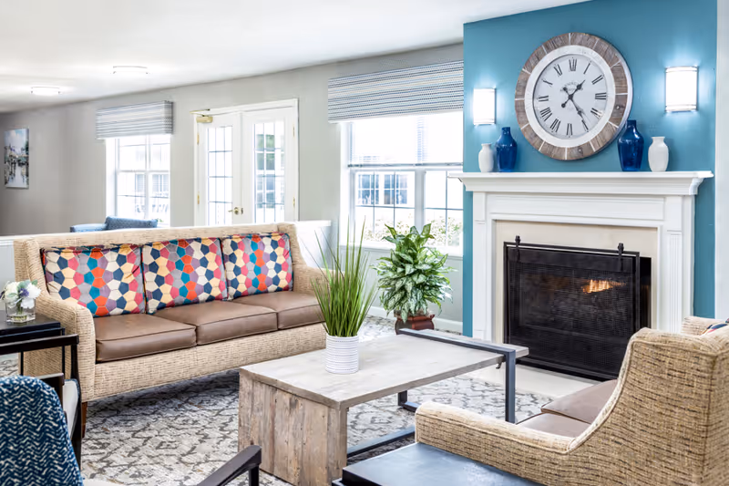 A bright and cozy living room with a beige sofa adorned with colorful geometric patterned cushions, a wooden coffee table with a potted plant, and a fireplace with a large round clock above it. The walls are painted in a combination of light gray and teal, and there are large windows letting in natural light.
