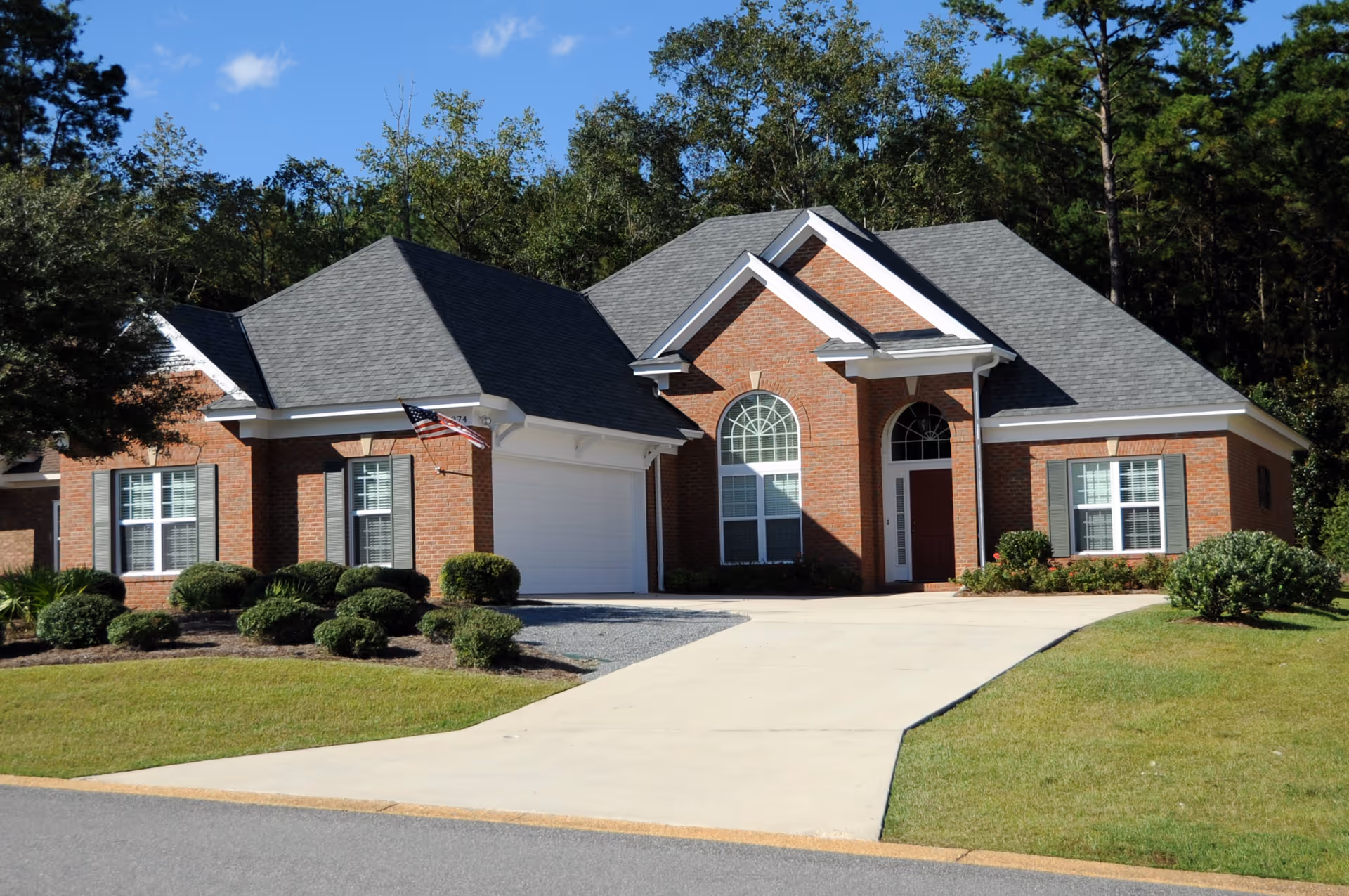 A single-story brick house with a gray shingled roof, white trim, and a two-car garage. The house features large arched windows and a red front door. The driveway is concrete and there are neatly trimmed bushes and a well-maintained lawn in front. Trees and a clear blue sky are visible in the background.