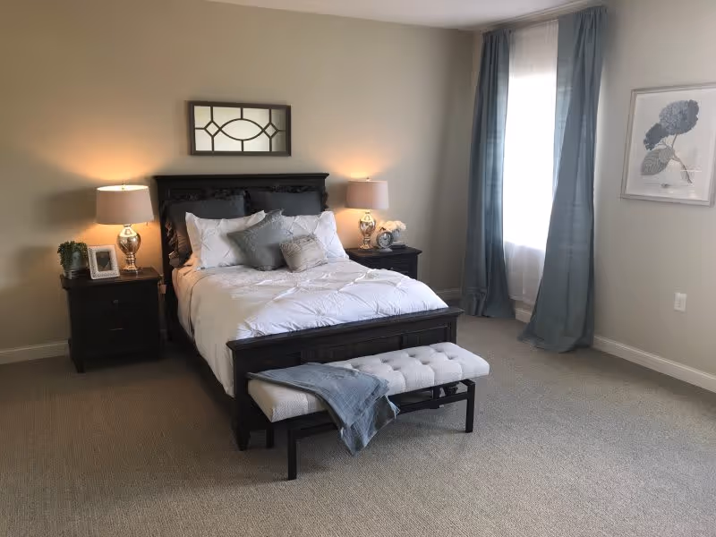 A neatly made bed with white bedding and multiple pillows in a bedroom. The bed has a dark wooden frame and is flanked by two matching nightstands, each with a lamp and decorative items. A cushioned bench with a folded blue throw is placed at the foot of the bed. A window with blue curtains and a framed botanical print are visible on the right wall.